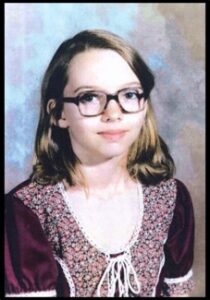 A young girl wearing glasses and a purple blouse poses for a picture, smiling slightly.